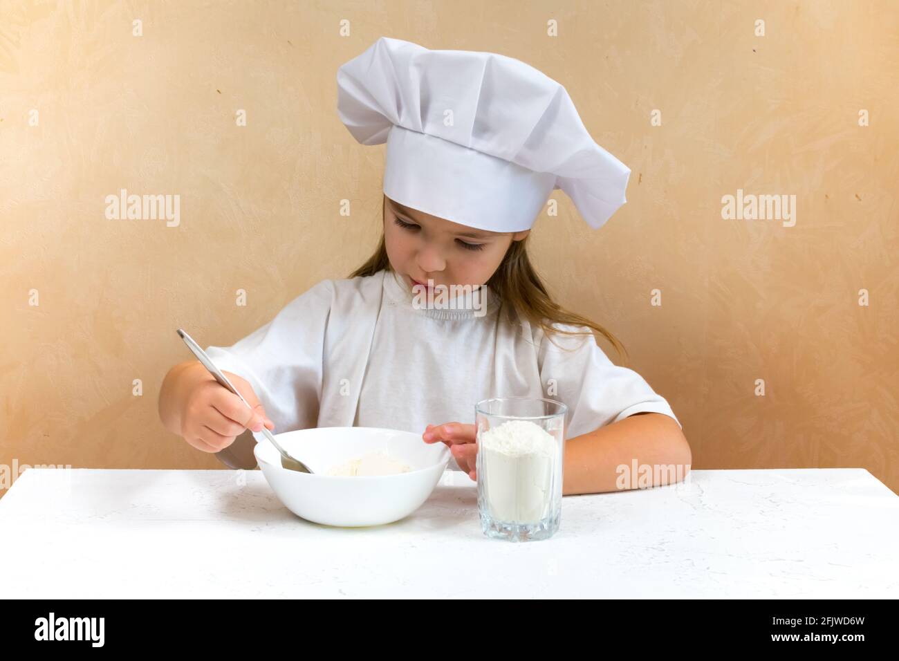 Una bambina vestita come cuoco impasta l'impasto. Cucina bambino stile di vita concetto. Il capretto ama, ha divertimento, studia e gioca nella cucina Foto Stock