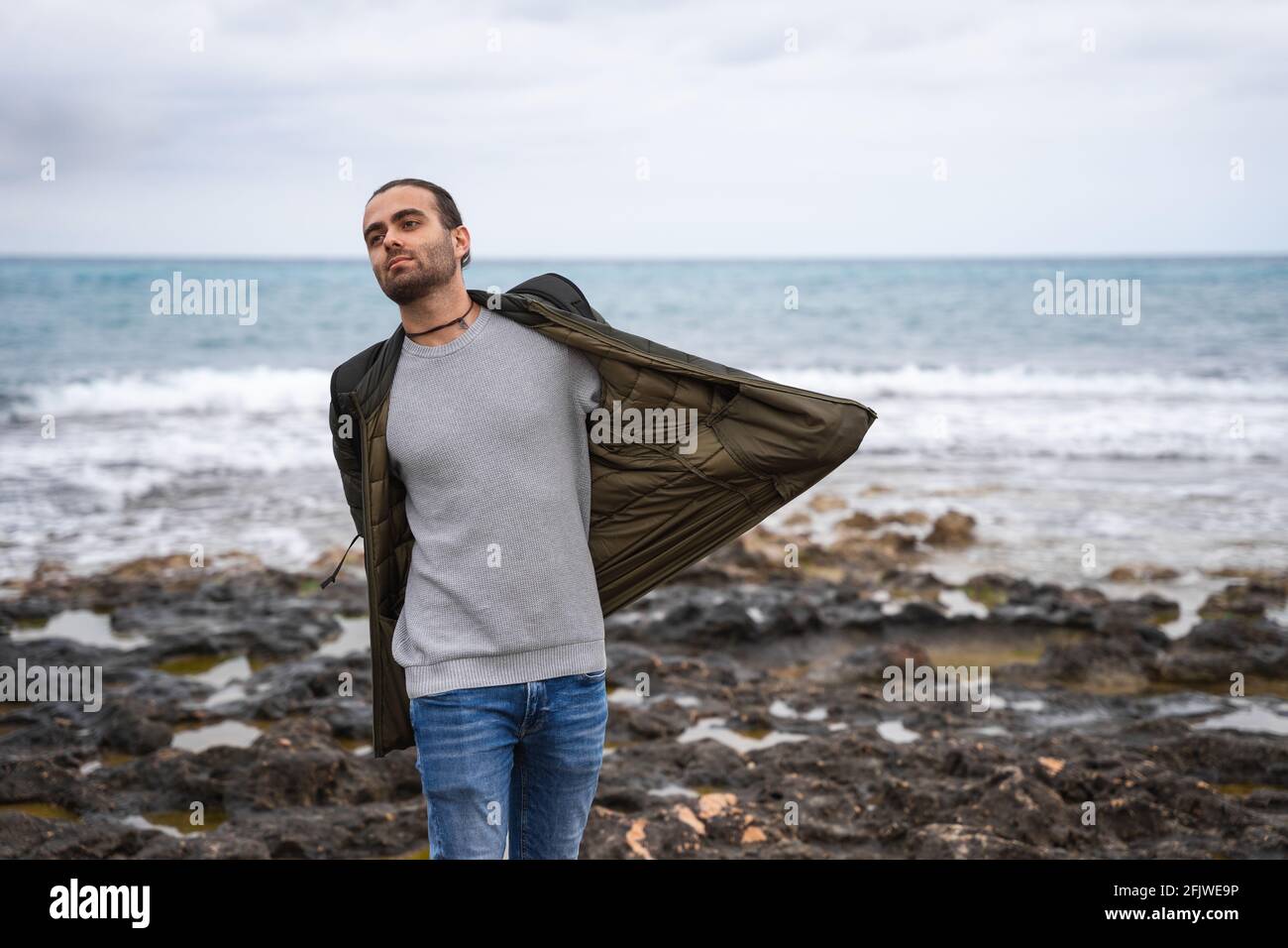 Uomo con le mani in tasca che stende la giacca. È in mare in un giorno nuvoloso. Indossa una giacca verde, un maglione grigio e jeans. Caucasica Foto Stock