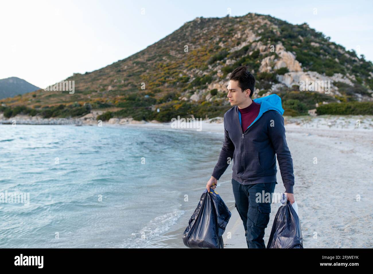 Giovane volontario uomo che cammina sulla spiaggia tenendo due sacchi pieni di rifiuti raccolti sul triste. Concetto di conservazione ambientale. Foto Stock