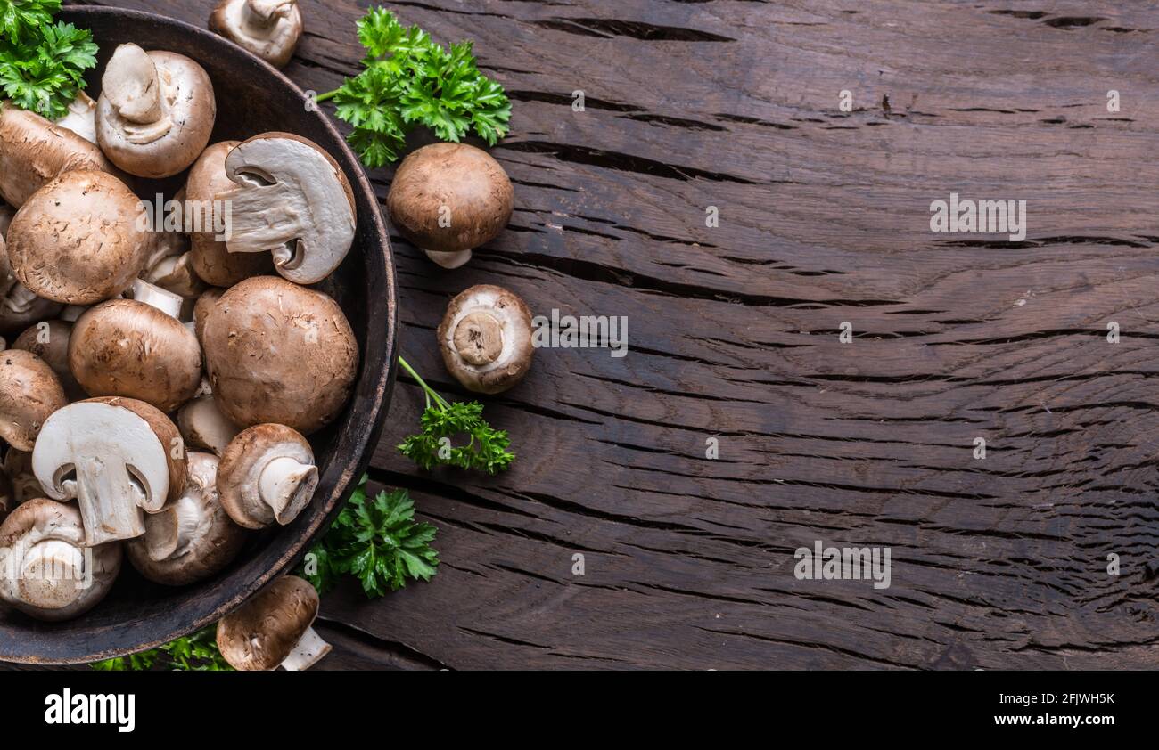 Funghi comuni di colore marrone in ciotola di legno su tavola di legno con erbe. Vista dall'alto. Foto Stock