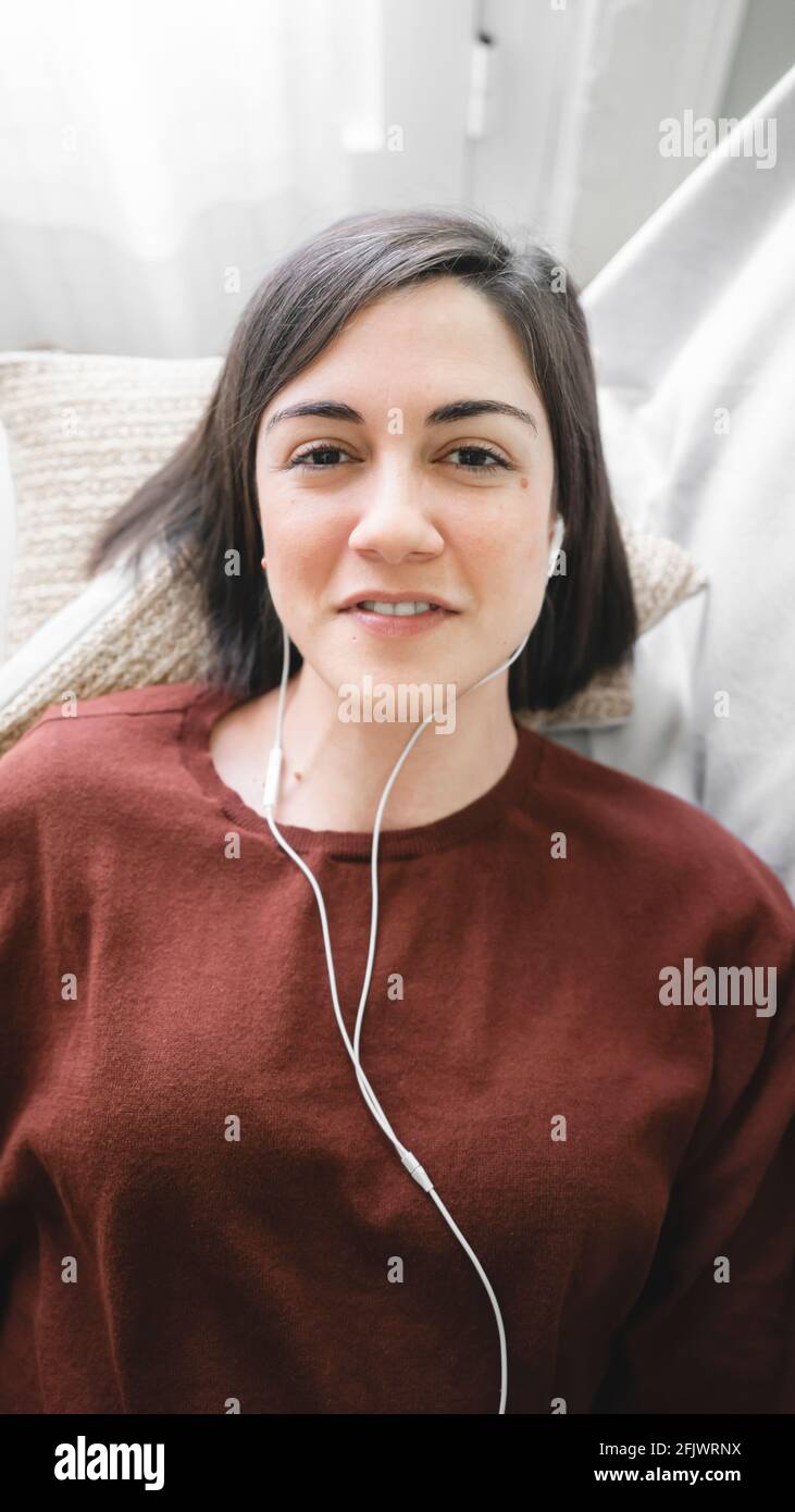 Donna sorridente con cuffie bianche che prende un selfie guardando la fotocamera si trova sul divano di casa Foto Stock