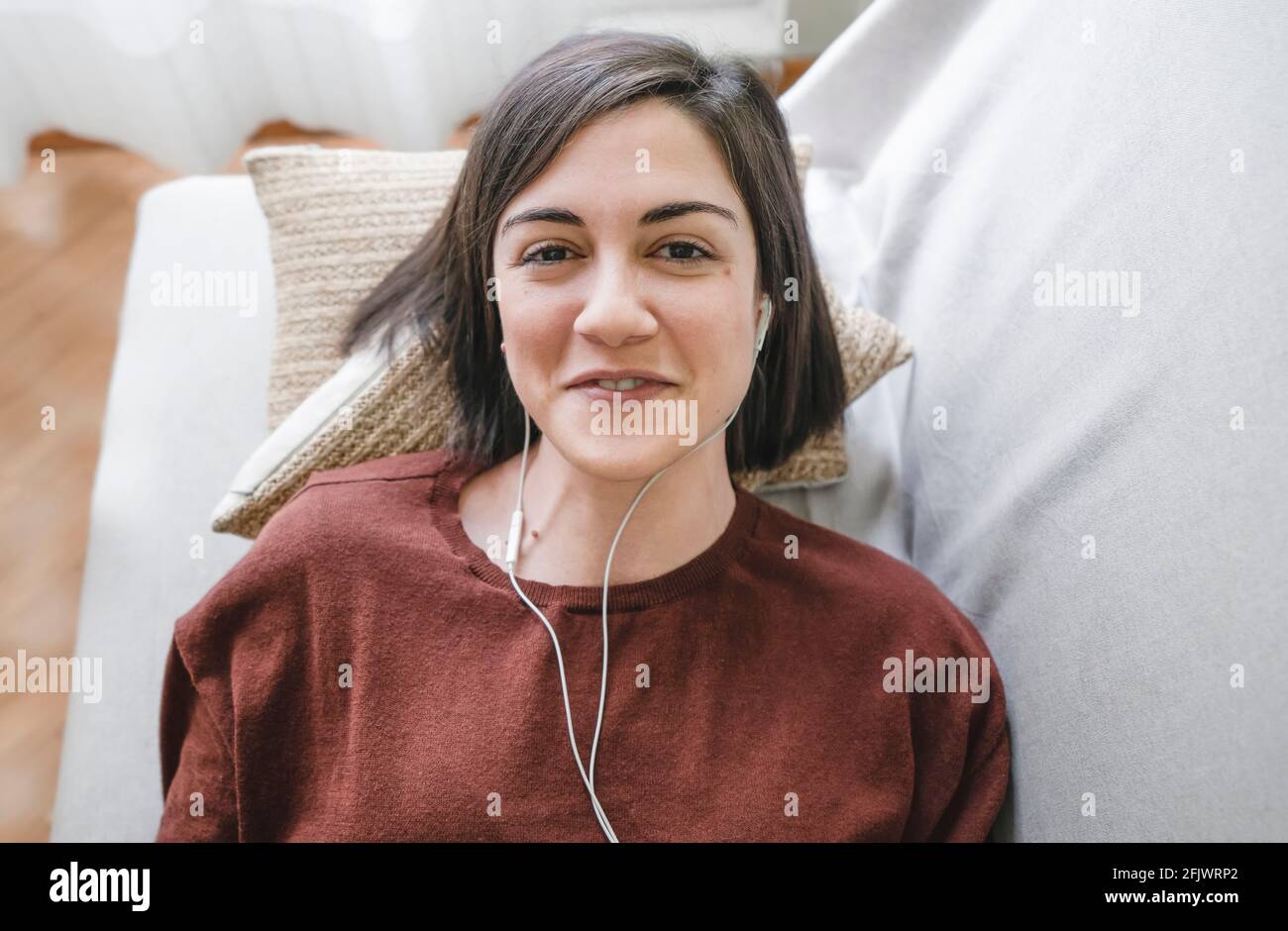 Donna sorridente con cuffie bianche che prende un selfie guardando la fotocamera si trova sul divano di casa Foto Stock