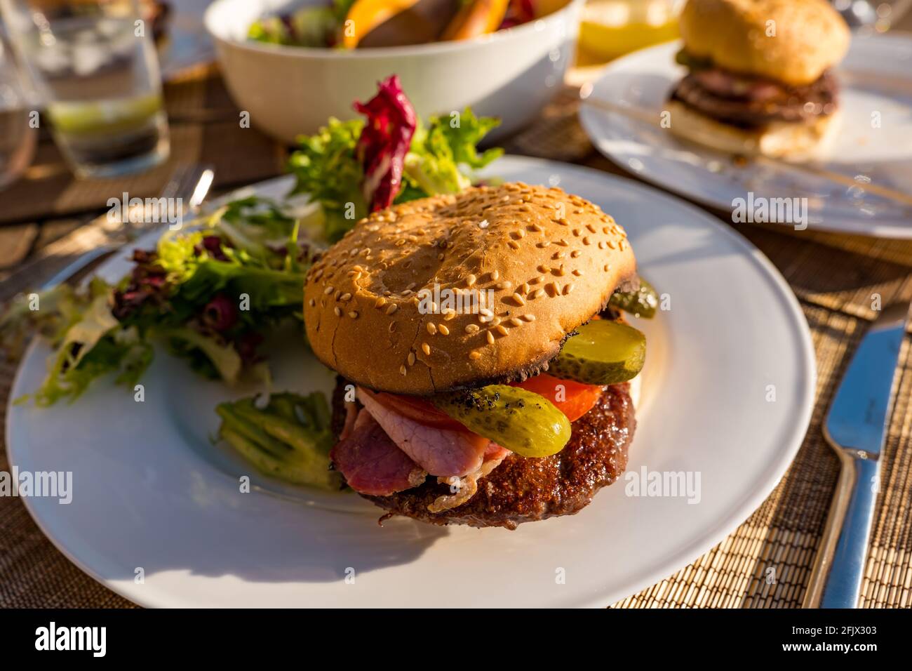 Hamburger di manzo con pancetta, pomodoro e sottaceto di aneto su un piatto bianco con insalata su un tavolo all'aperto sotto il sole Foto Stock