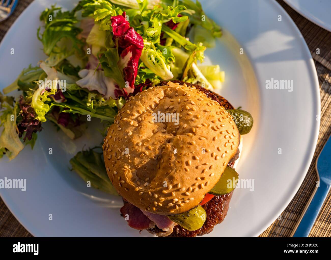 Hamburger di manzo con pancetta, pomodoro e sottaceto di aneto su un piatto bianco con insalata su un tavolo all'aperto sotto il sole Foto Stock