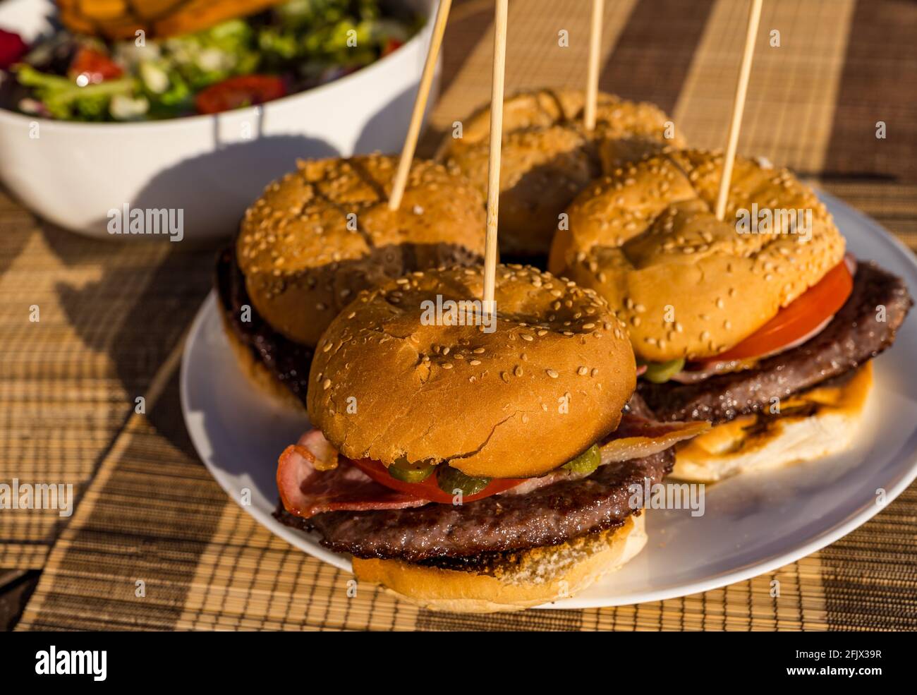 Hamburger di manzo con pancetta, pomodoro e sottaceto di aneto sul piatto con recipiente per insalate all'aperto sotto il sole Foto Stock