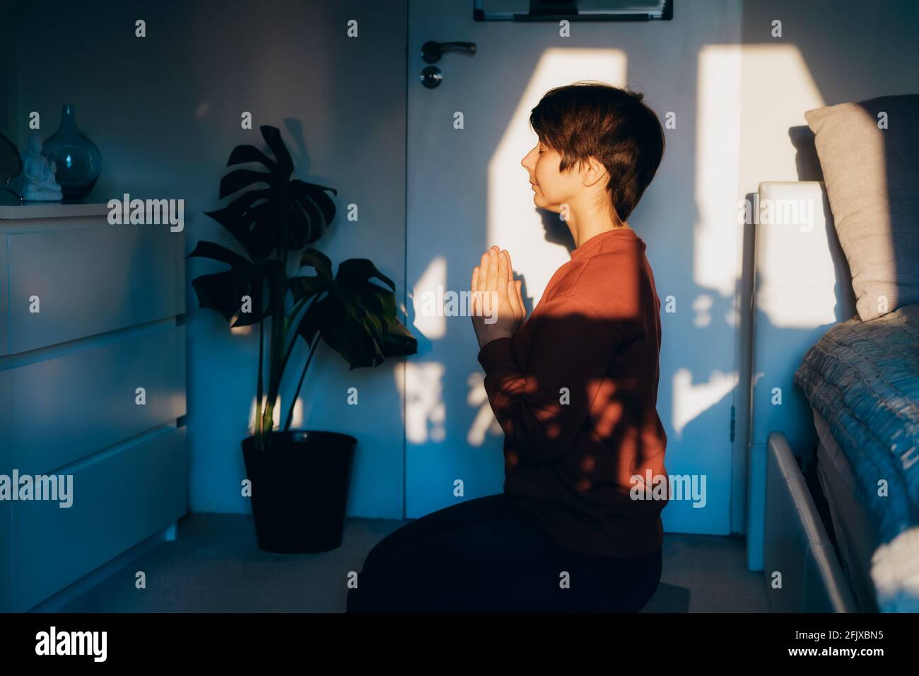 Donna con occhi chiusi seduta su un tappetino in camera da letto, facendo Namaste nella luce del tramonto. Rituale di preghiera con fede alla statua del buddha a casa. Zen Foto Stock