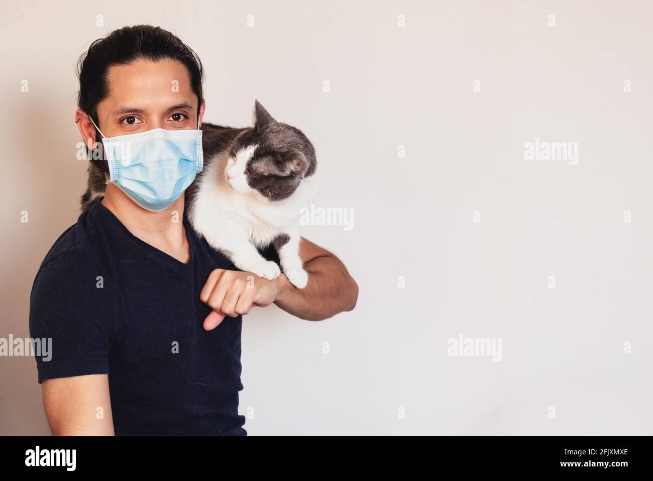 Giovane latino con un gatto sulle spalle che indossa Una maschera per la sicurezza da COVID-19 su uno sfondo chiaro con spazio per la copia Foto Stock