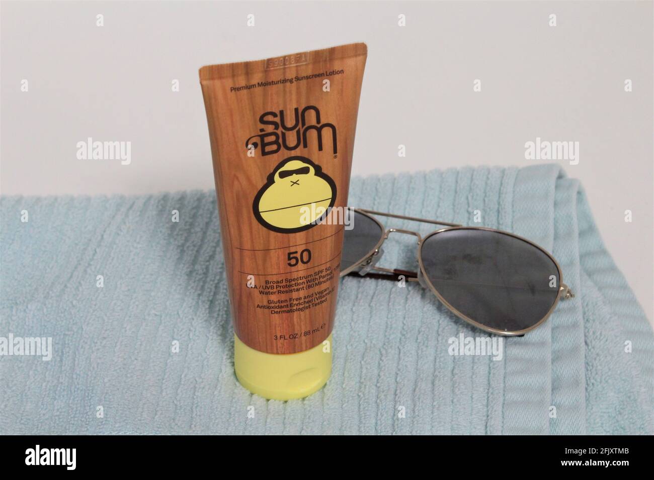 Crema solare Sun Bum visualizzata su un asciugamano blu accanto agli occhiali da sole su sfondo bianco. Foto Stock
