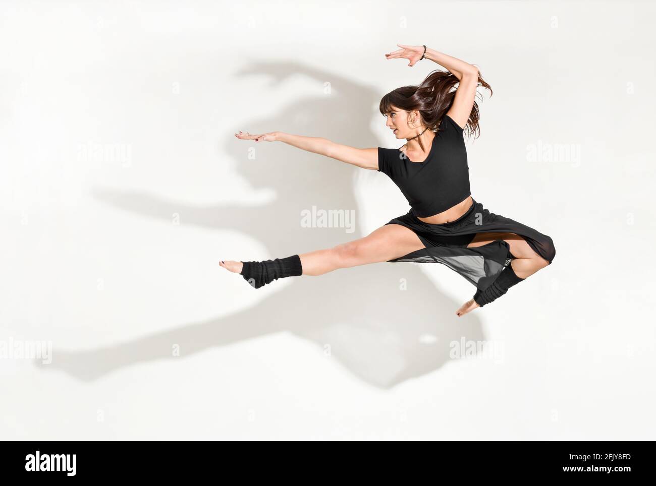 Giovane ballerina agile che esegue un salto frontale semi-diviso con i capelli lunghi che volano in una posa a mezz'aria isolata sopra bianco con ombra drammatica e copyspac Foto Stock