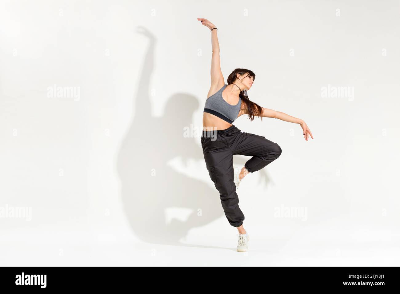 Aggraziata giovane donna danzatrice in un hip hop outfit esecuzione una classica posa da ballo con braccia allungate che si bilanciano su una gamba con ombra artistica su a. Foto Stock