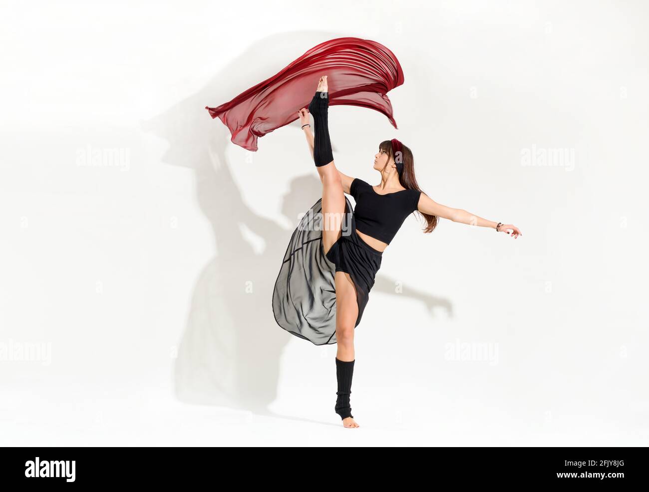 Giovane ballerina agile che esegue una posizione dinamica di pattinaggio su ghiaccio alzando la gamba in aria come lei galleggia con grazia tessuto rosso sopra la sua testa isola Foto Stock