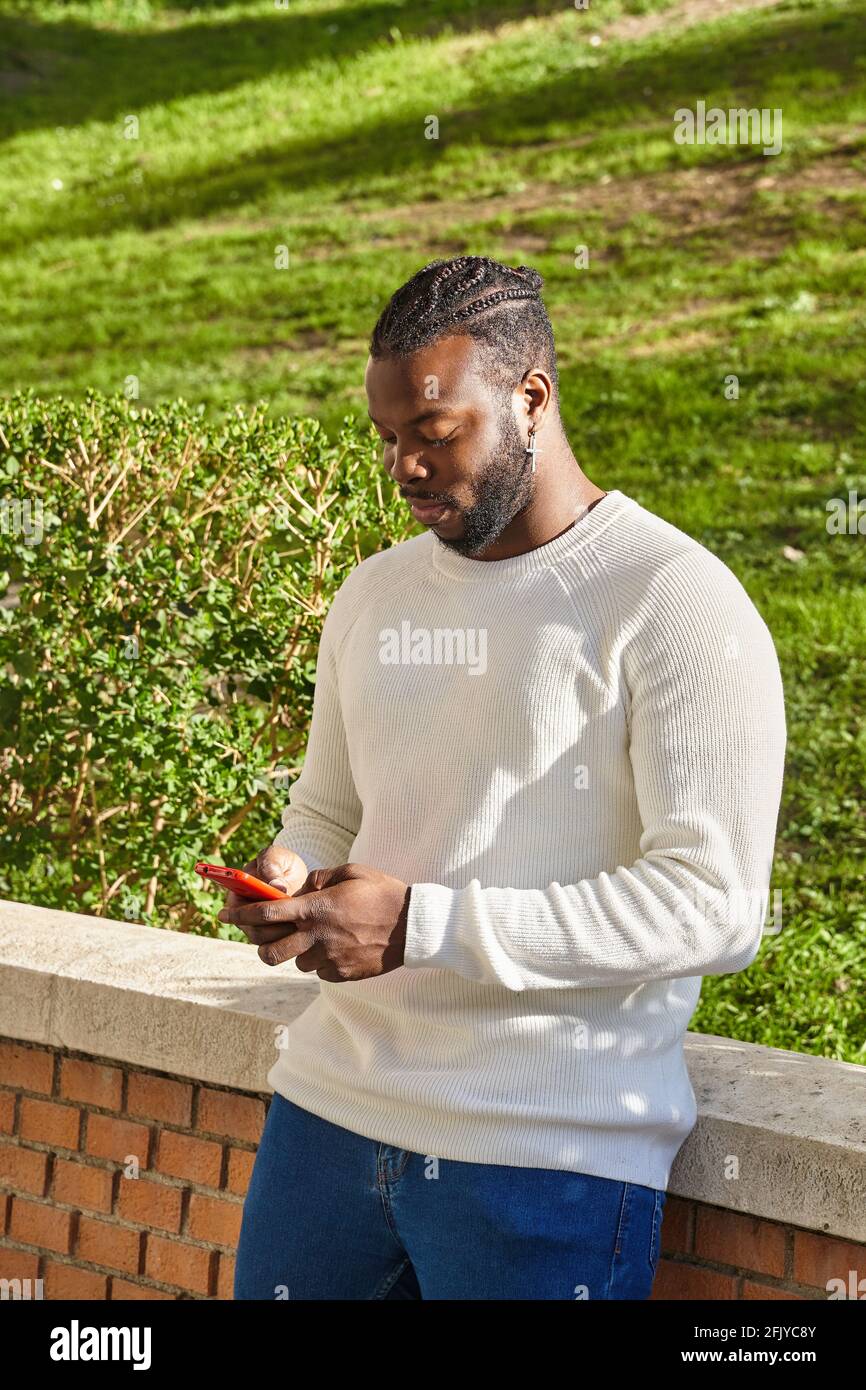 Bell'uomo afro-americano che chiacchiera tramite smartphone in piedi, allegro uomo dalla pelle scura godendo di tempo libero blogging in reti tramite smartphone. Foto di alta qualità Foto Stock