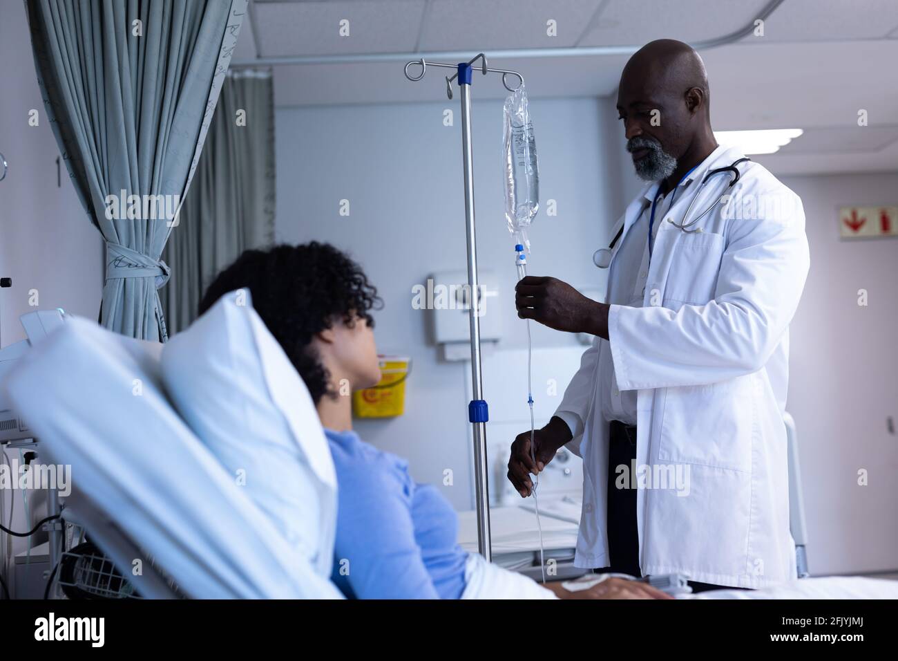 African american maschio medico preparante iv sacchetto di gocciolamento per mescolato donna di razza seduta in letto d'ospedale Foto Stock