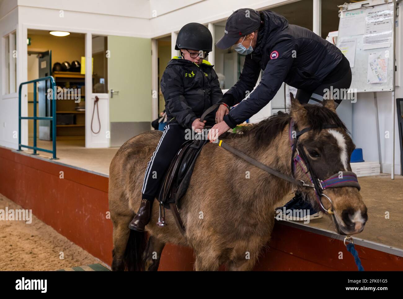 East Lothian, Scozia, Regno Unito, 27 aprile 2021. Riapre Muirfield Riding Therapy: La carità accoglie i suoi primi giovani per la loro sessione di terapia con restrizioni di blocco attenuati. A causa delle restrizioni in corso, al momento possono essere soddisfatti solo i clienti più mobili. Nella foto: Fin, di 10 anni, un allievo alla scuola primaria di yester, è aiutato nella sua terapia cavallo Briony da sua madre Foto Stock