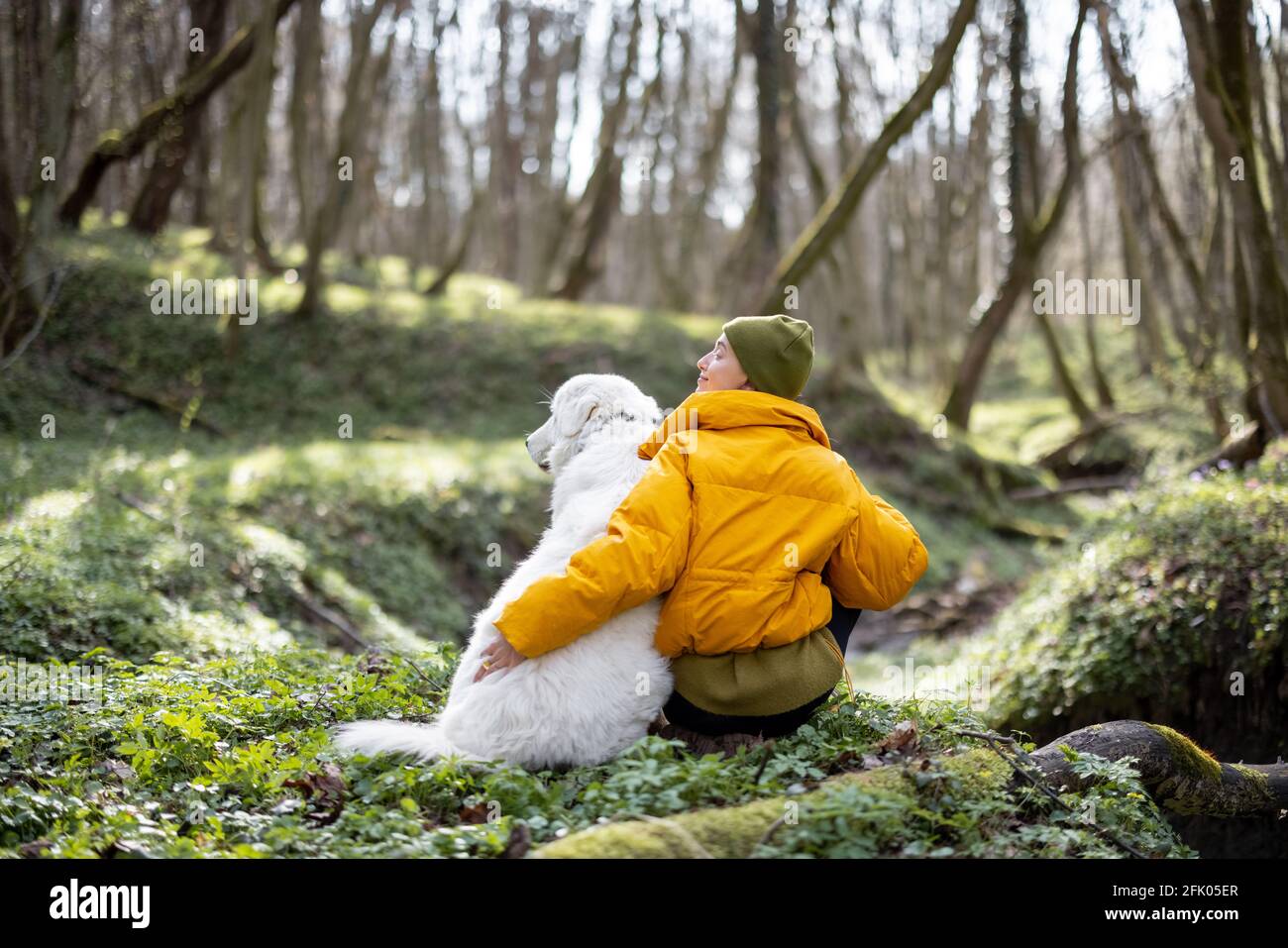 Giovane donna in abiti da trekking seduto vicino al grande cane bianco in verde primavera foresta. Gode ed esplora la natura tranquilla. Foto Stock