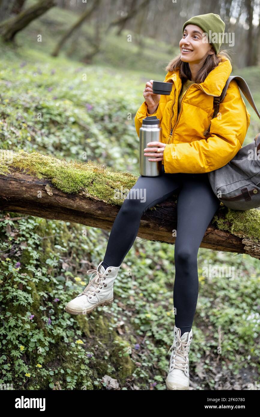 Giovane donna in abiti colorati e zaino da trekking seduta su un albero e bere tè caldo da un thermos. Gode della freschezza dell'aria e della tranquillità della natura nella foresta primaverile. Foto Stock