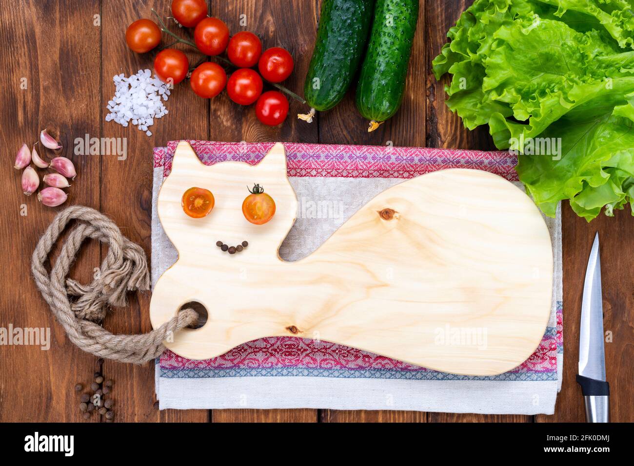 Taglio di tavole di legno sotto forma di gatto su un tavolo di legno. Occhi di pomodoro di ciliegia e un sorriso di pepe. Tagliere fatto a mano. Pomodori freschi, cucum Foto Stock