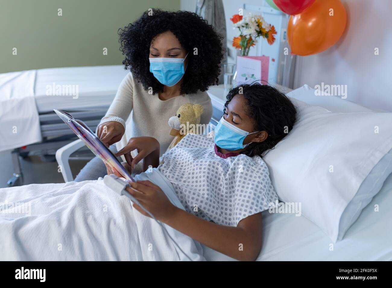 Razza mista madre e figlia malata in maschere di viso in ospedale, libro di lettura con orsacchiotto Foto Stock