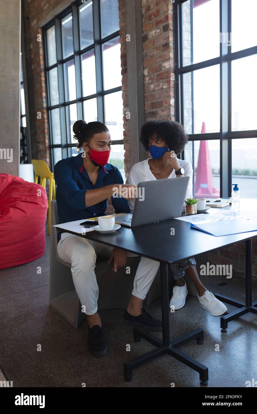 Donna afroamericana e uomo da corsa misto che indossa maschere facciali sedersi in un bar utilizzando un computer portatile Foto Stock