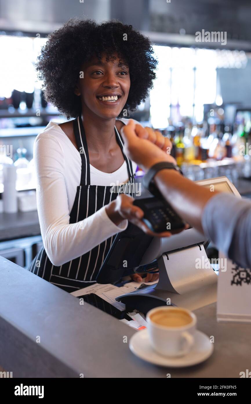 Sorridente barista afroamericana con grembiule, usando il terminale di pagamento Foto Stock