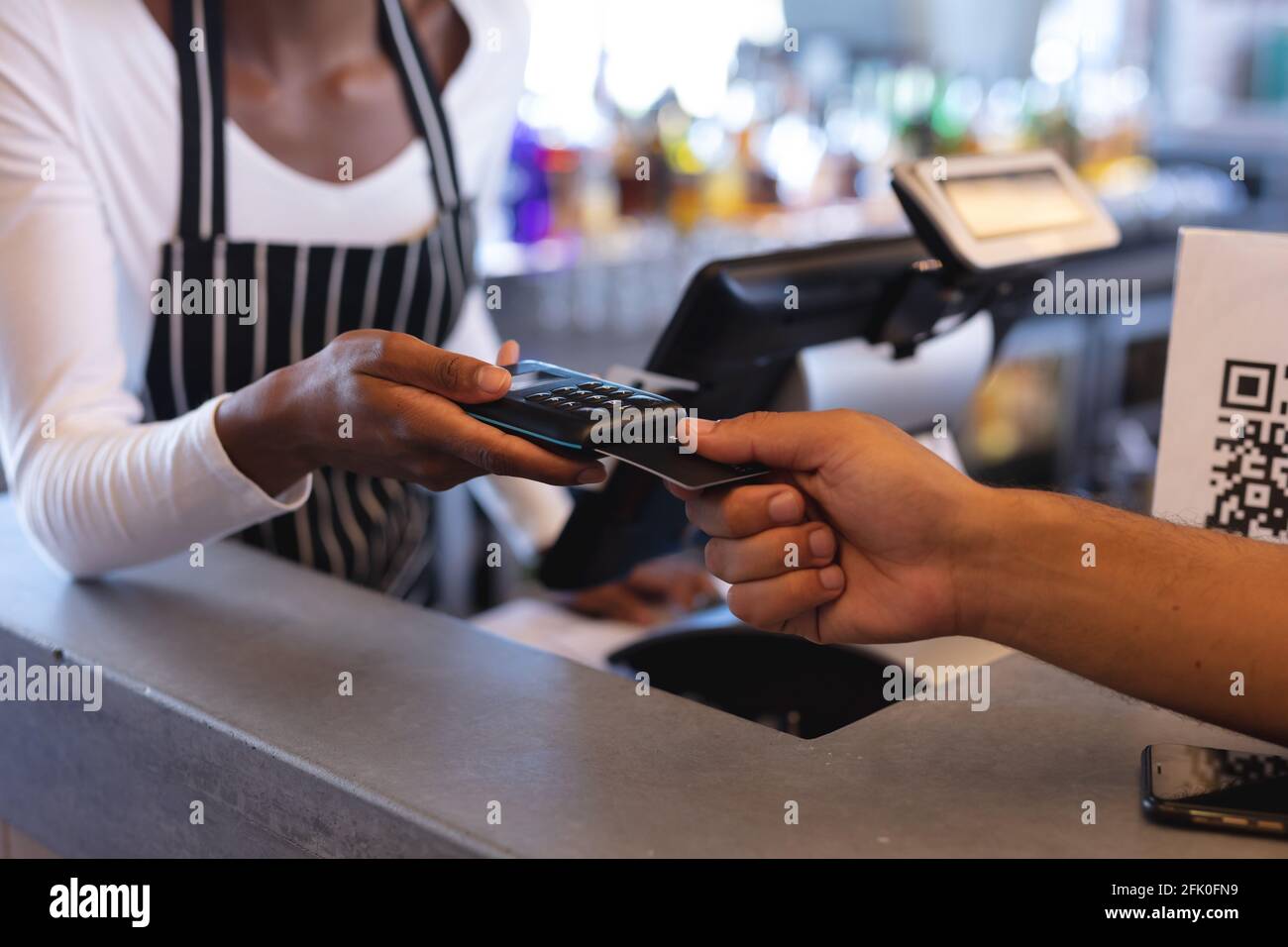 Sezione centrale della cameriera afroamericana che indossa un grembiule, usando il terminale di pagamento nel caffè Foto Stock