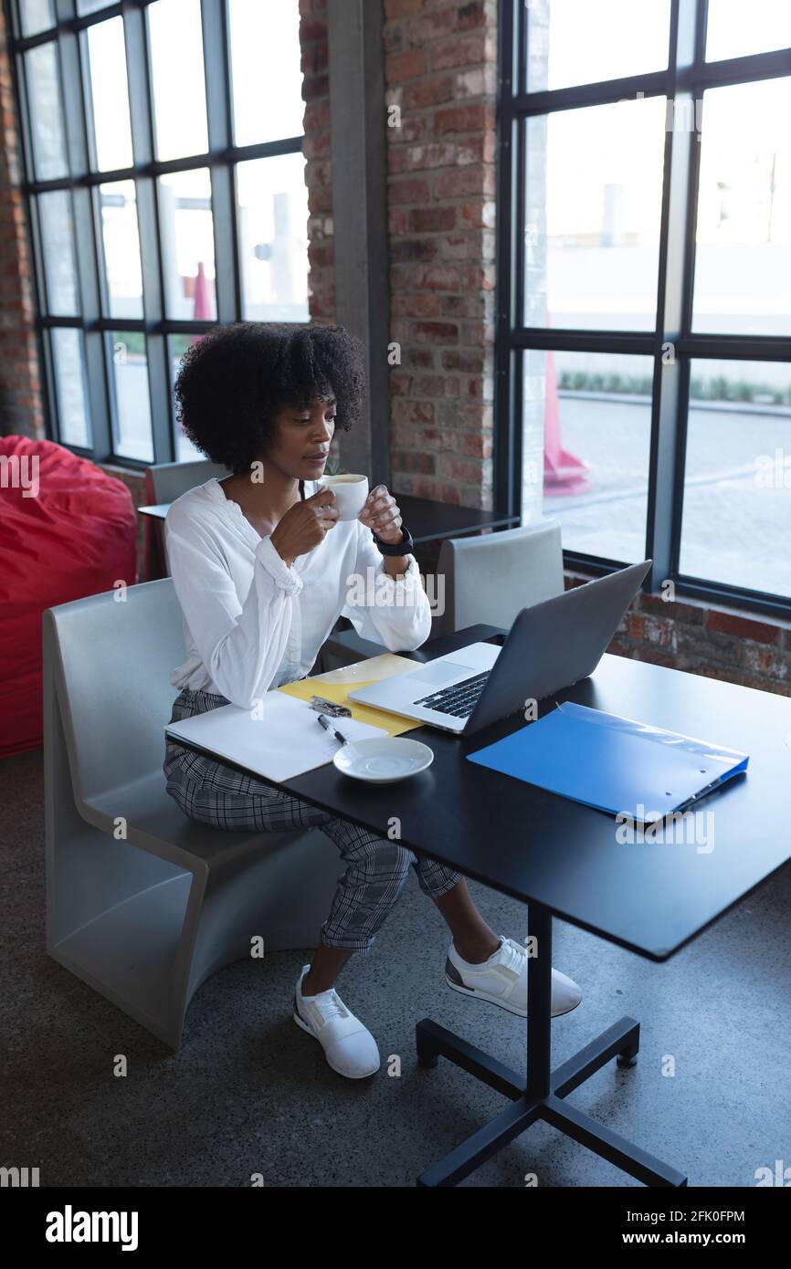 Donna afroamericana che beve caffè, utilizzando il computer portatile e lavorando in un bar Foto Stock