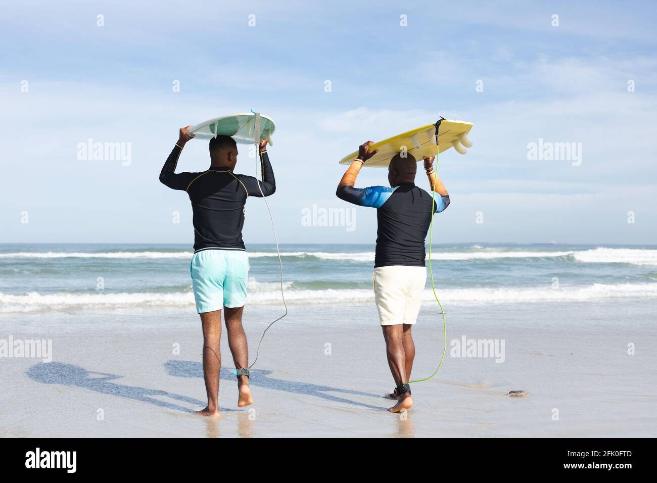 Vista posteriore del padre e del figlio afro-americani che trasportano tavole da surf sulla loro testa alla spiaggia Foto Stock