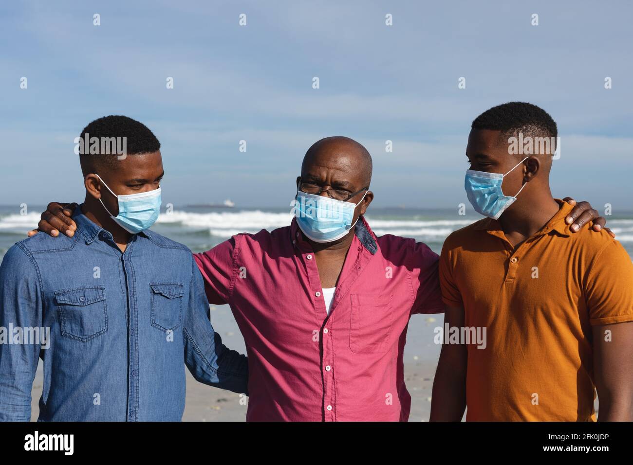 Il padre afroamericano e i suoi due figli indossano maschere facciali stare insieme in spiaggia Foto Stock