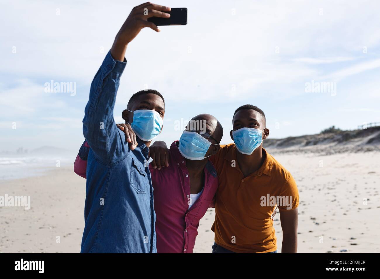Il padre afroamericano e i suoi due figli indossano maschere facciali prendere un selfie insieme alla spiaggia Foto Stock