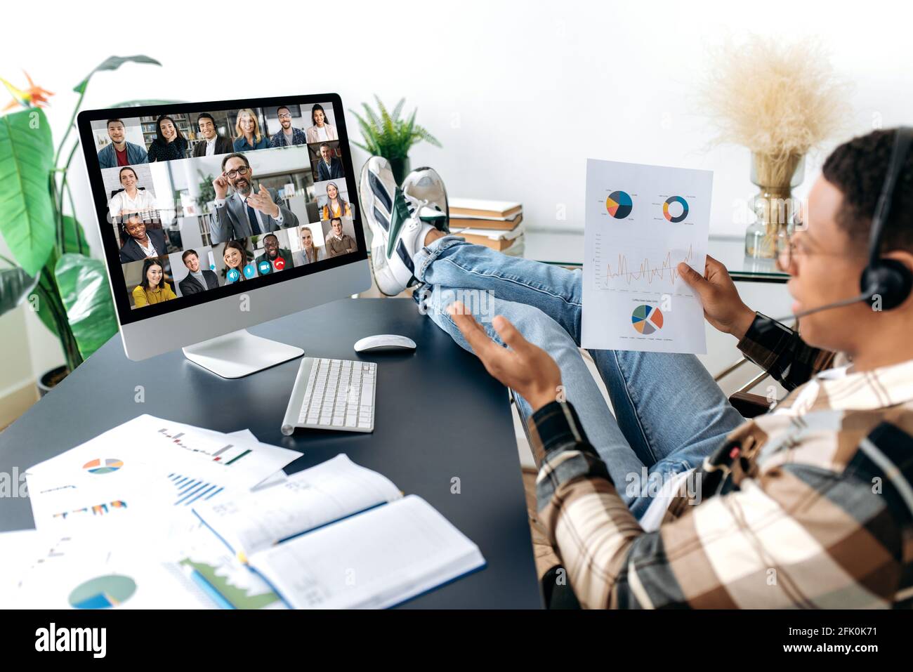 Videoconferenza, riunione online. L'uomo d'affari intelligente di razza mista si siede sul posto di lavoro, con i piedi gettanti sul tavolo, con le cuffie, comunica con la videochiamata con i suoi partner, discute una strategia Foto Stock