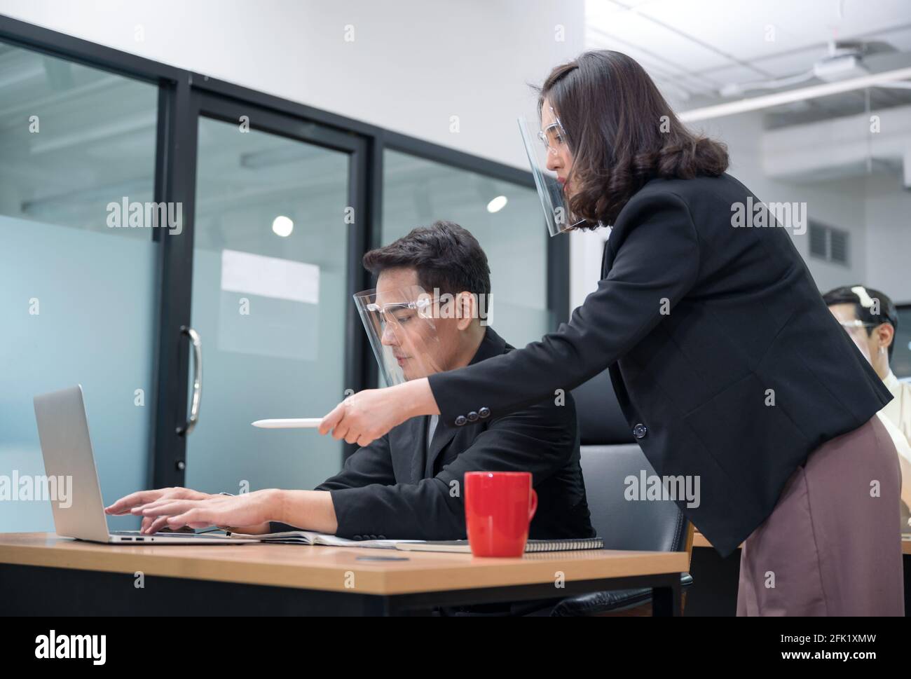 Bossy manager femmina ordinare e puntare al portatile di ufficio lavoratore maschile che indossa la visiera di protezione in un nuovo ufficio normale Foto Stock