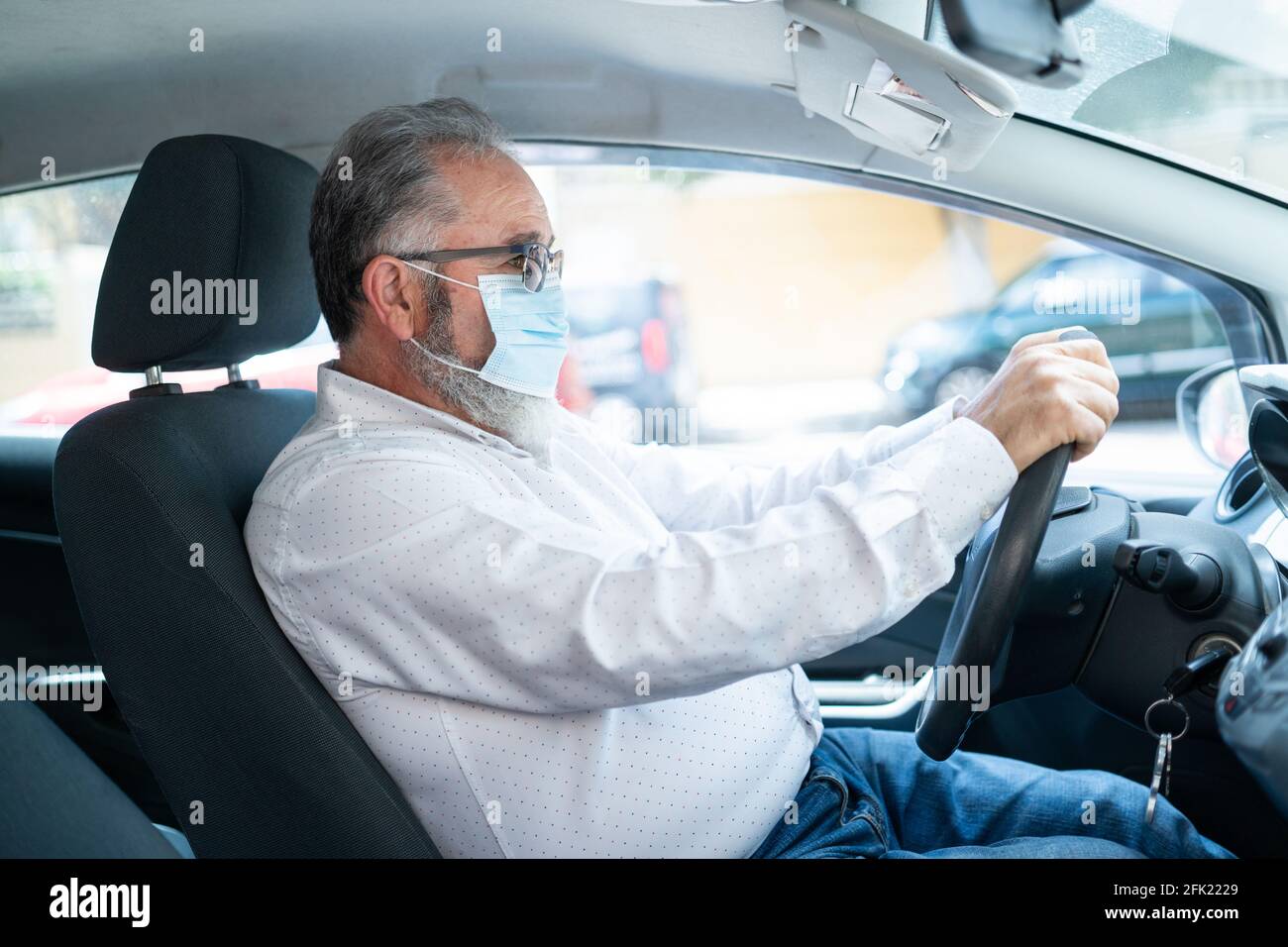 vista laterale di un uomo anziano in una maschera chirurgica medica che guida un'automobile. Concetto pandemico di coronavirus. Foto Stock
