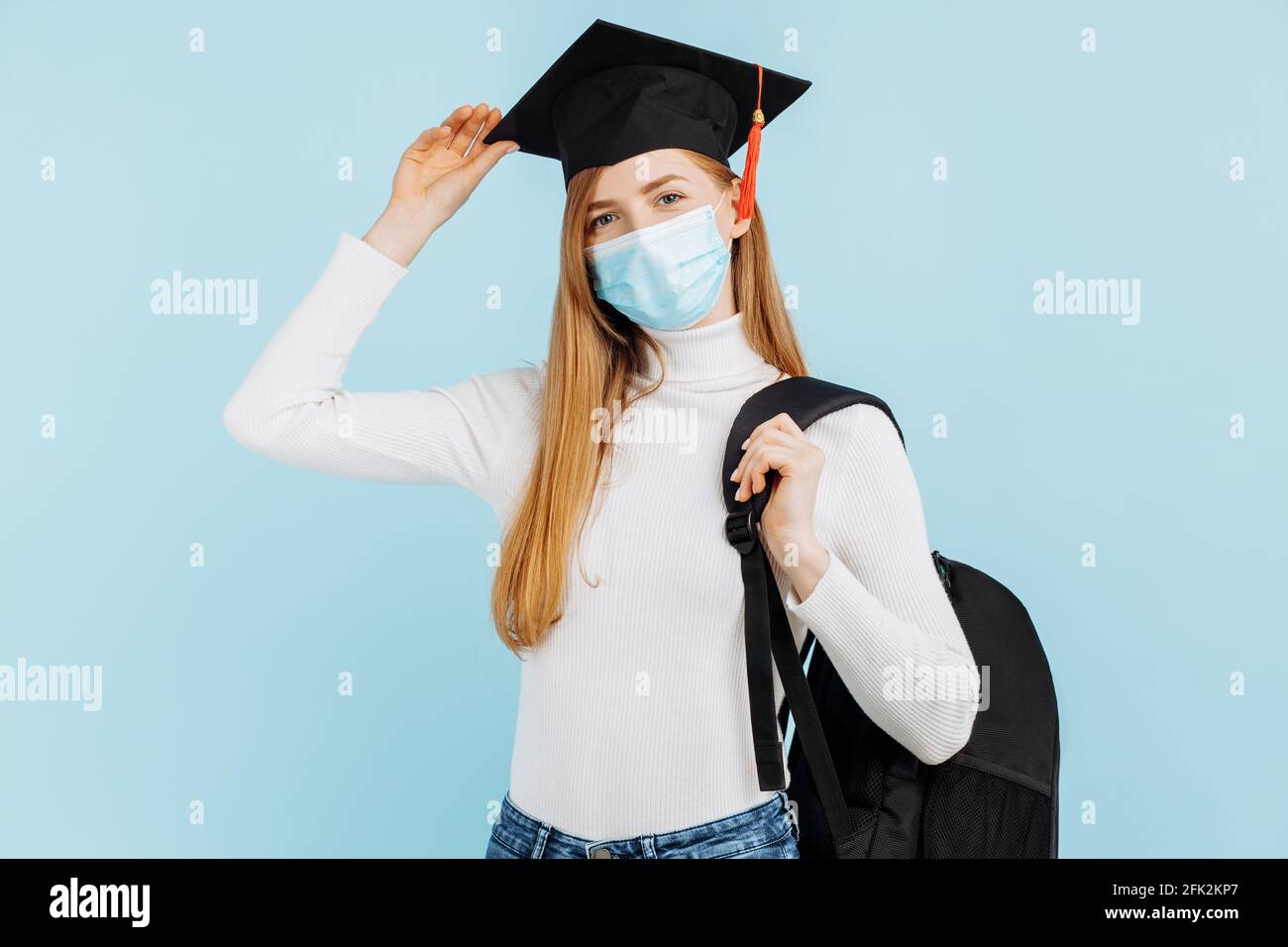 Felice giovane donna laureata in maschera medica, studente in cappello di laurea con valigetta su sfondo blu Foto Stock