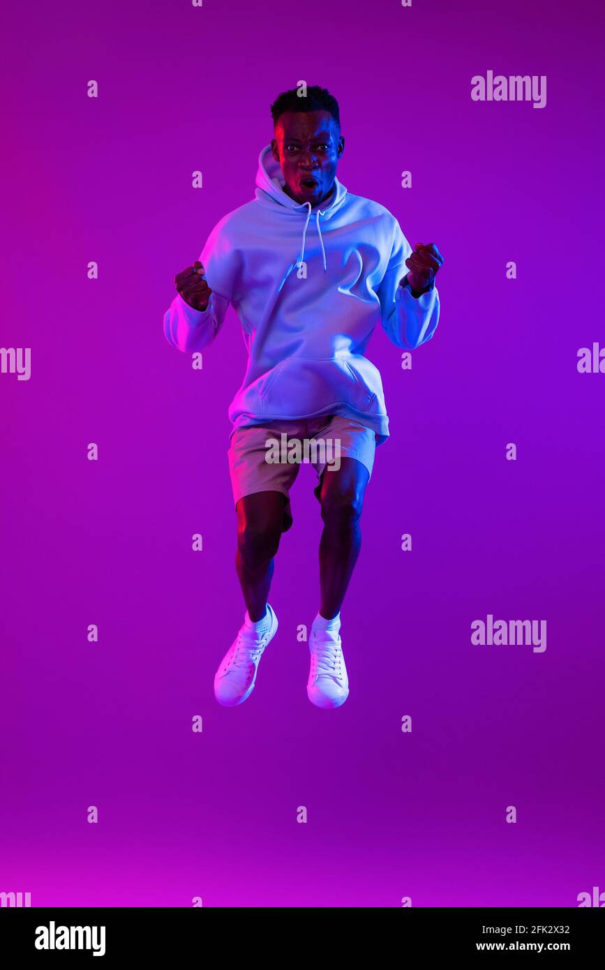 Giovane uomo africano estatico che salta in futuristico cyberpunk viola neon sfondo chiaro Foto Stock