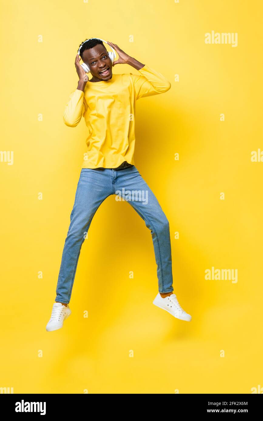 Ritratto a tutta lunghezza di un giovane africano felice che indossa le cuffie ascoltare musica e saltare in uno sfondo giallo isolato studio Foto Stock