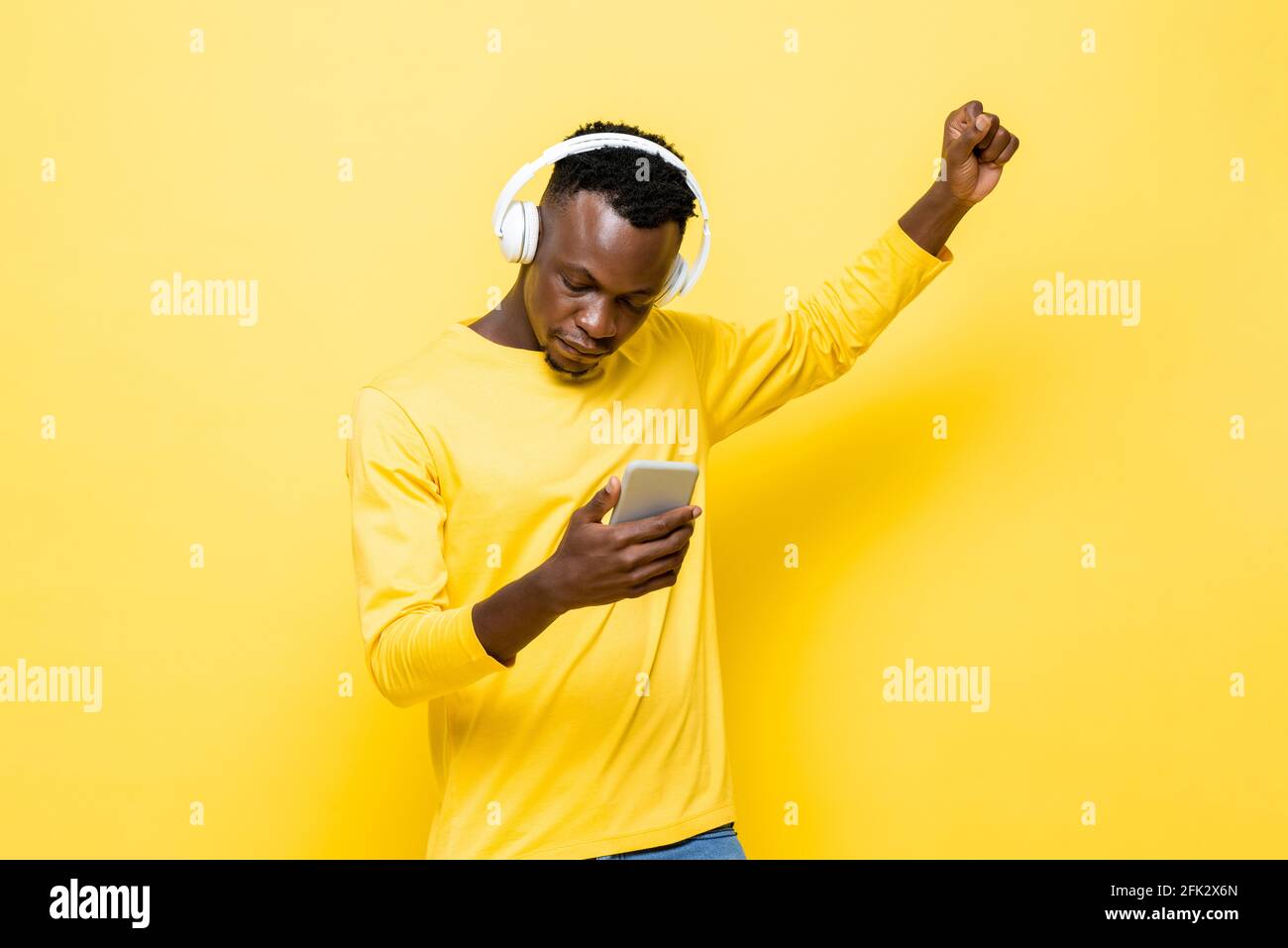 Giovane uomo africano in abbigliamento casual con cuffie senza fili in ascolto alla musica con il telefono cellulare su sfondo giallo isolato studio Foto Stock
