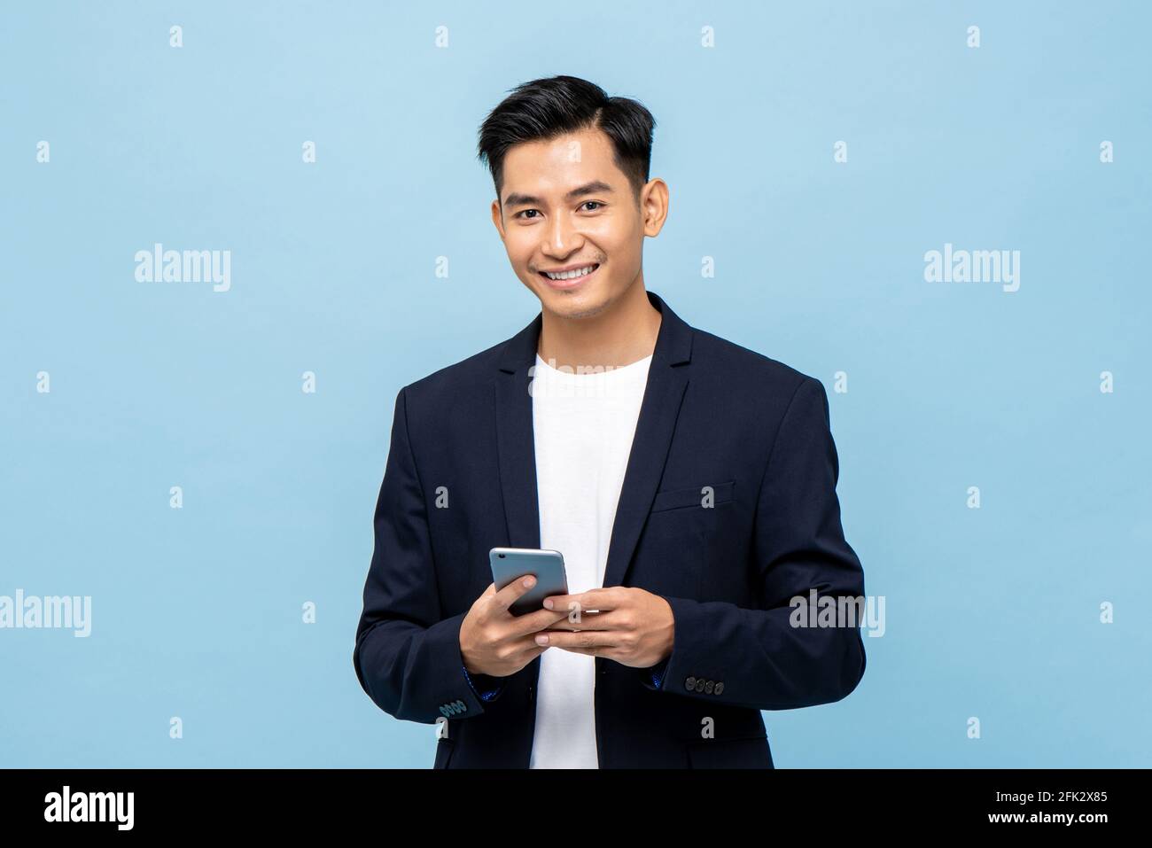Vita in su ritratto di giovane sorridente bell'uomo asiatico dentro tuta semi-formale con telefono cellulare in blu chiaro isolato sfondo studio Foto Stock