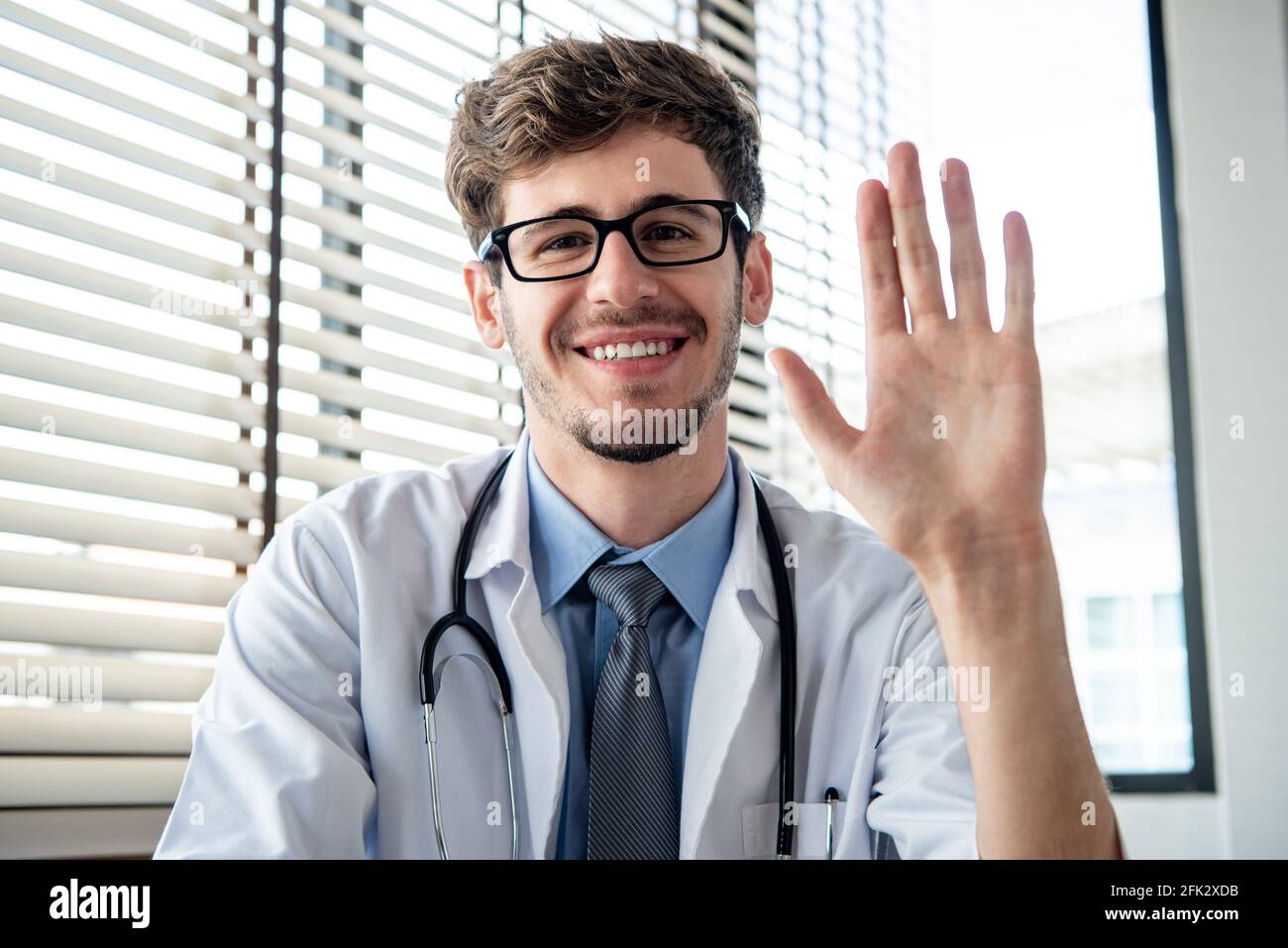 Sorridente giovane medico maschio che agitava la mano alla macchina fotografica salutando il paziente online tramite videochiamate a distanza dall'ufficio Foto Stock