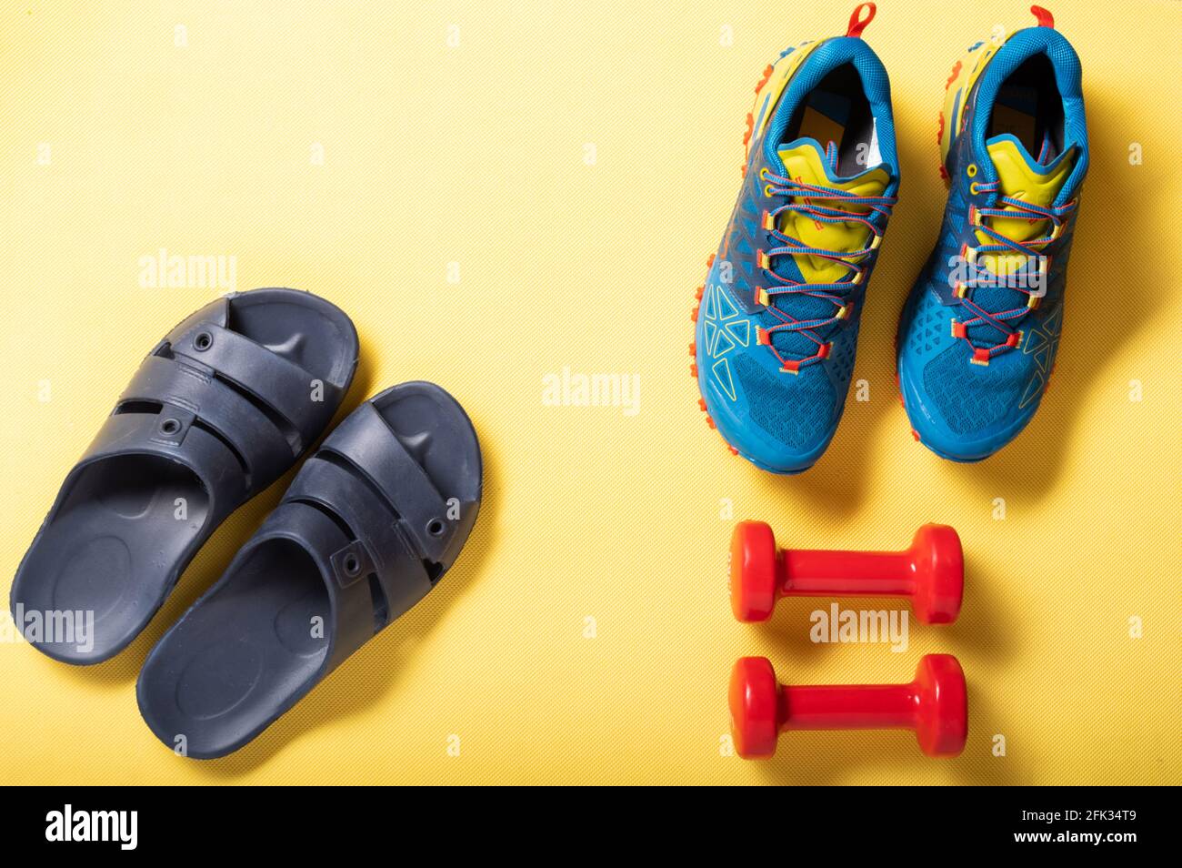 Ciabatte per la casa e scarpe sportive. Scarpe sportive e manubri su un tappetino giallo. In alto. Foto Stock