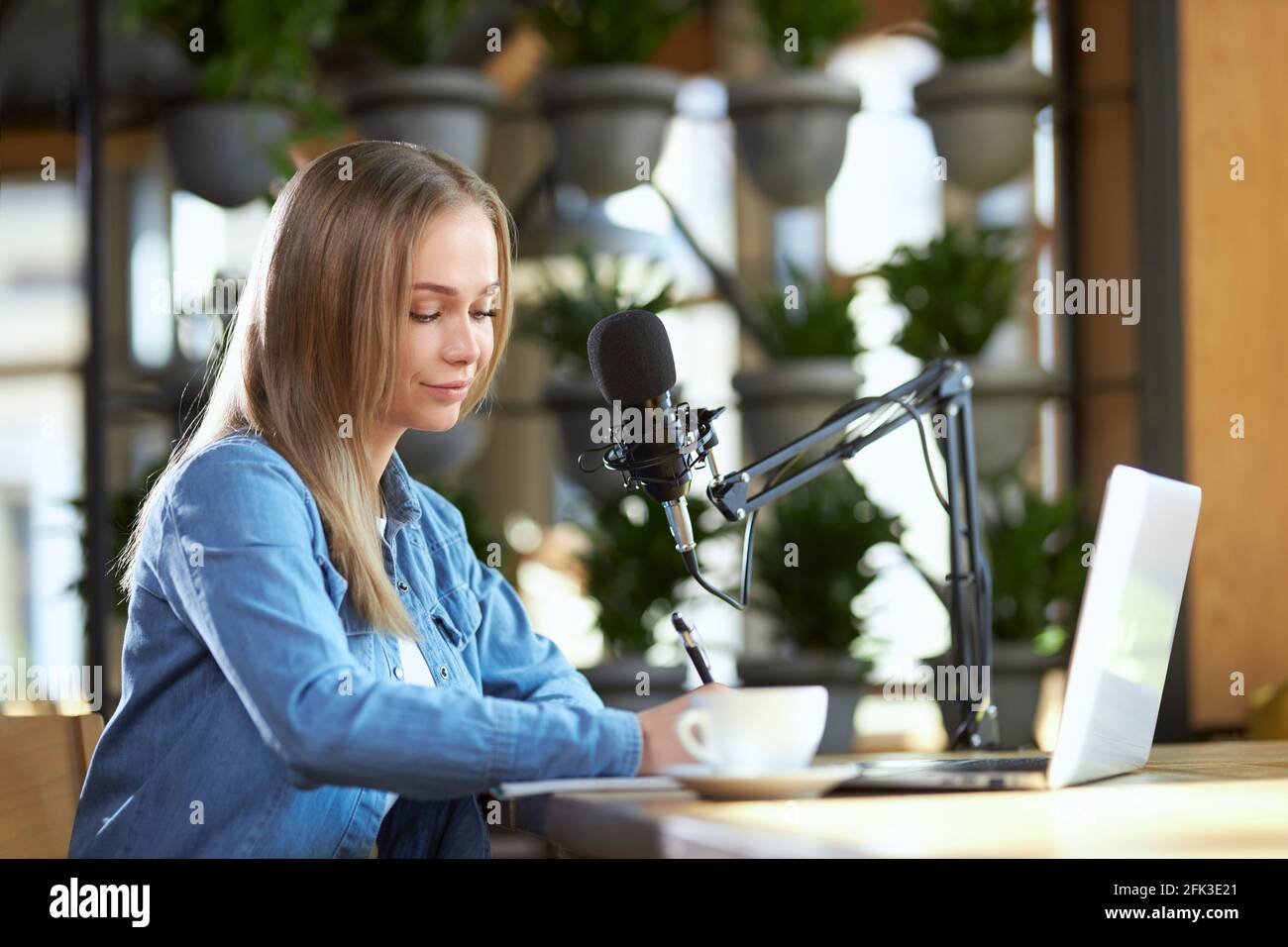 Vista laterale di una giovane donna sorridente attraente in giacca denim seduta e scrittura di informazioni in notebook. Concetto di processo preparazione trasmissione live con caffè gustoso. Foto Stock