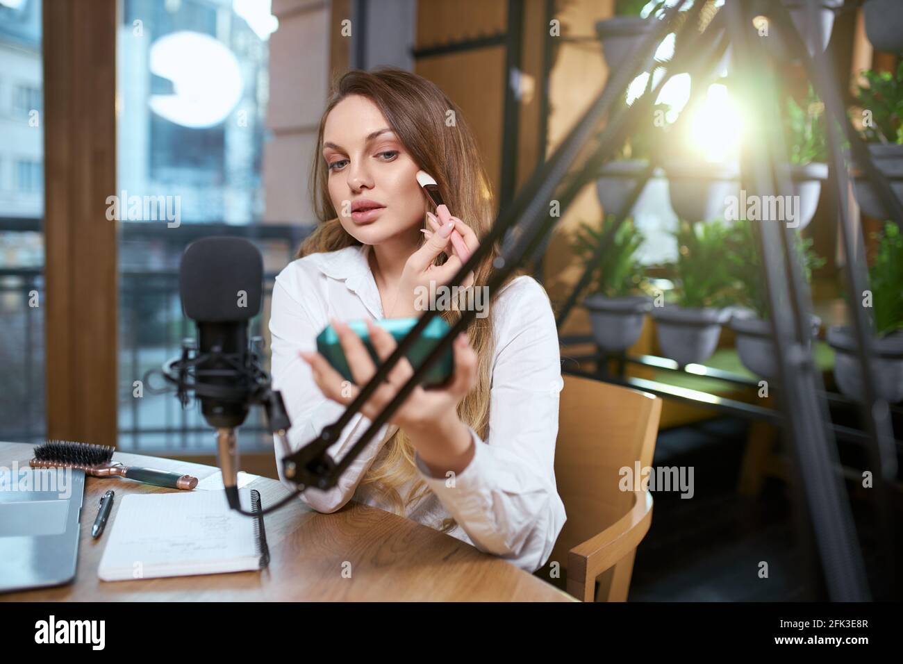 Primo piano ritratto di giovane bella donna in camicia bianca facendo il trucco vicino ad attrezzature musicali speciali in caffetteria. Concetto di processo preparazione per l'intervista in trasmissione online. Foto Stock