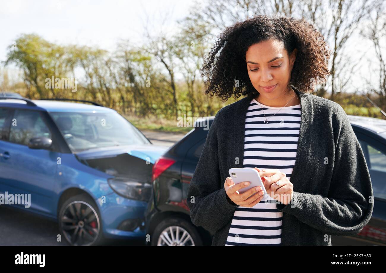 Giovane donna in piedi da auto danneggiata dopo la segnalazione di incidente stradale incidente con compagnia assicurativa che utilizza il telefono cellulare Foto Stock
