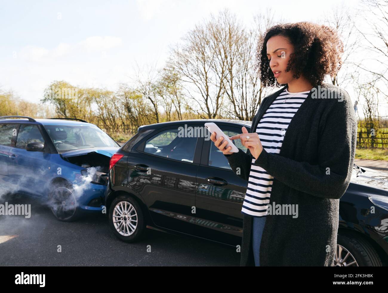 Giovane donna in piedi da auto danneggiata dopo la segnalazione di incidente stradale incidente con compagnia assicurativa che utilizza il telefono cellulare Foto Stock