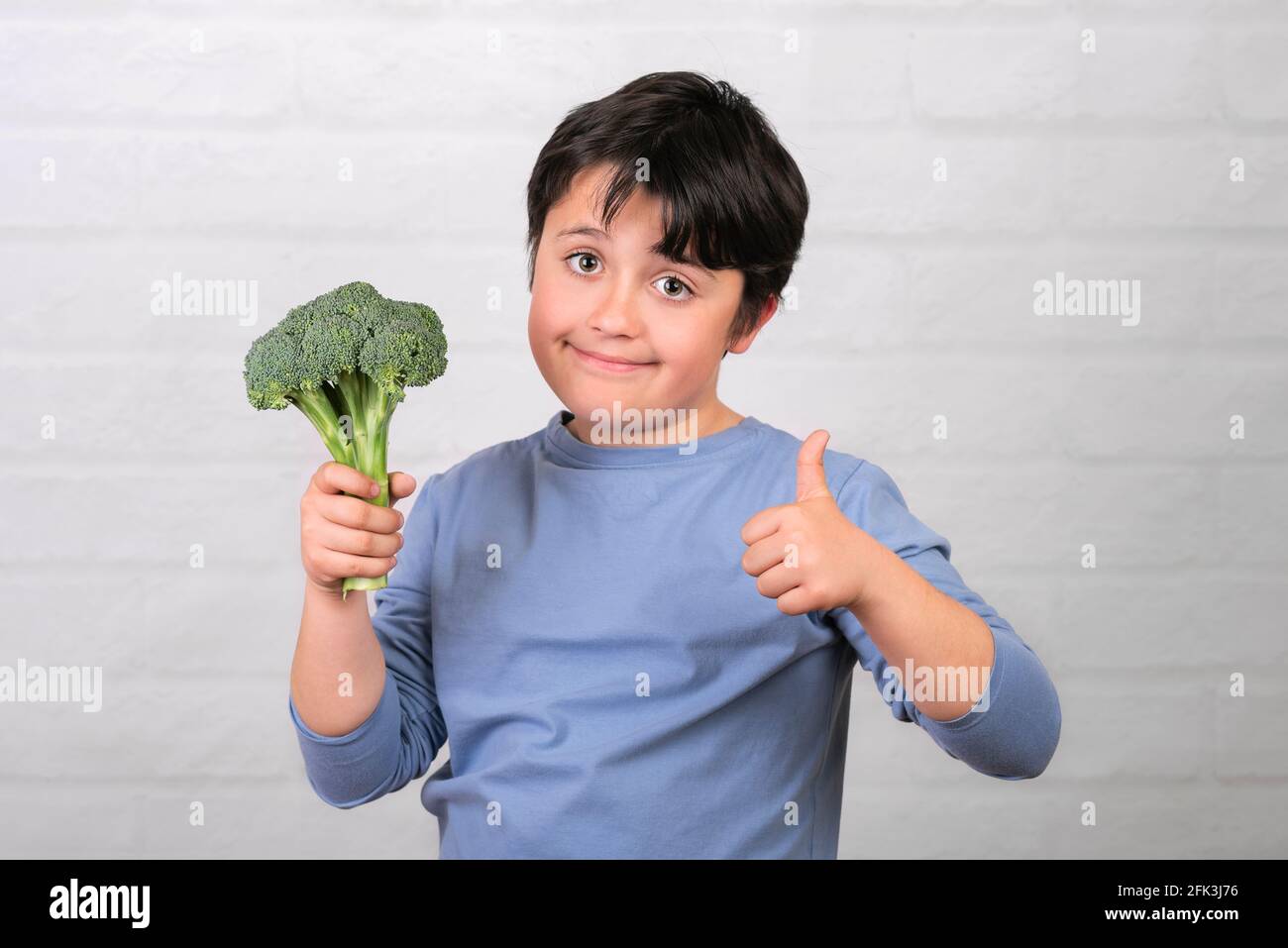 Bambino felice con broccoli in mano mostrando il pollice in su Su muro bianco di mattoni background.concetto di cibo sano Foto Stock