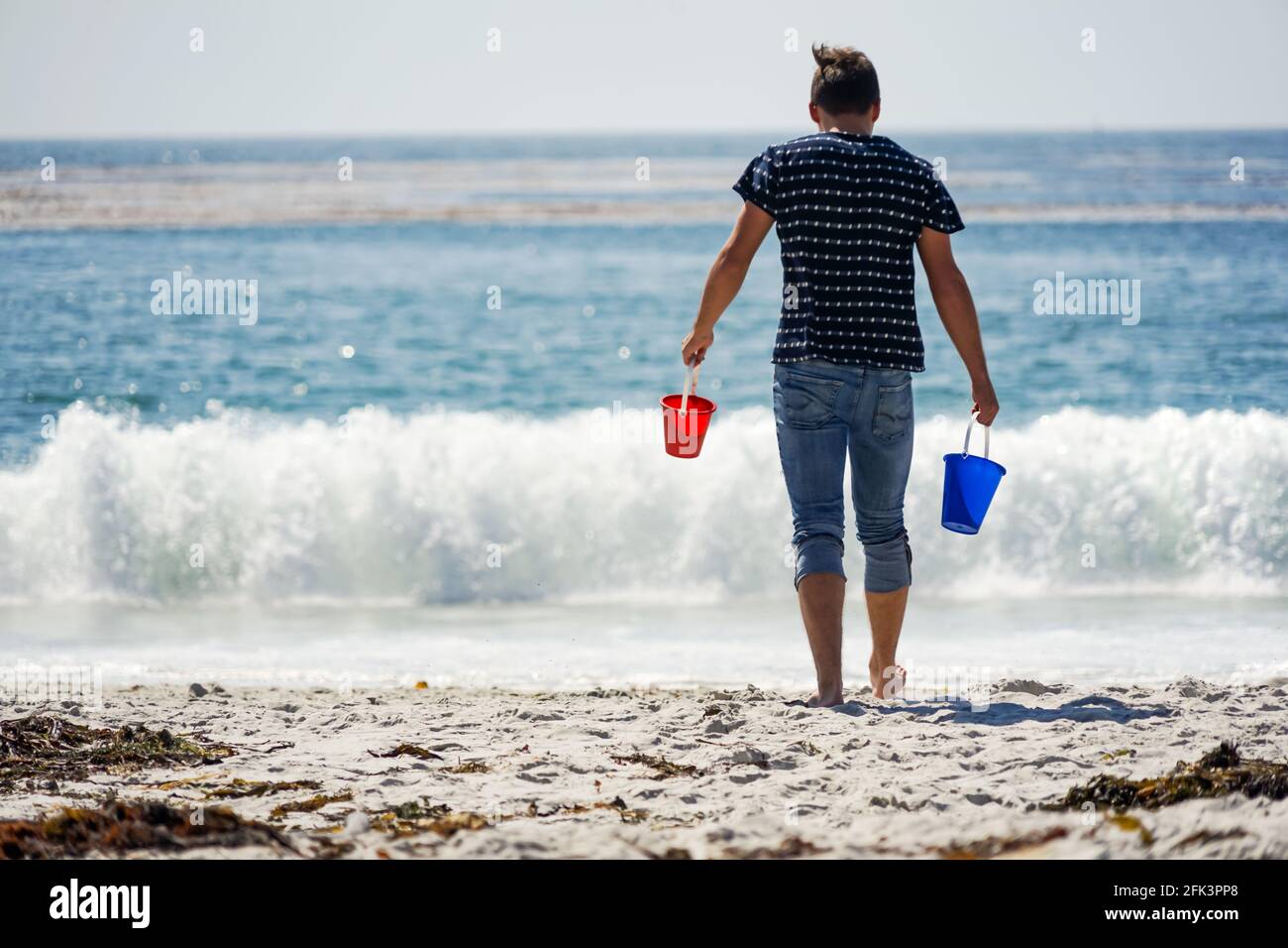 Un giovane uomo su una spiaggia soleggiato che si incetta acqua per il suo castello di sabbia in due piccole secchielli rosso e blu Foto Stock