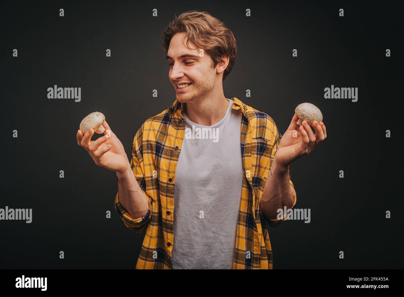 Il giovane biondo in camicia gialla tiene i funghi in entrambe le mani, isolato su sfondo nero Foto Stock