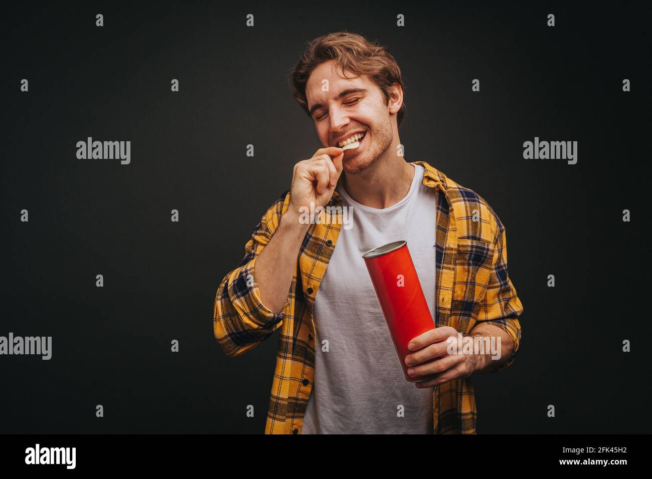 Giovane biondo uomo in camicia gialla sta mangiando chip, isolato su sfondo nero Foto Stock