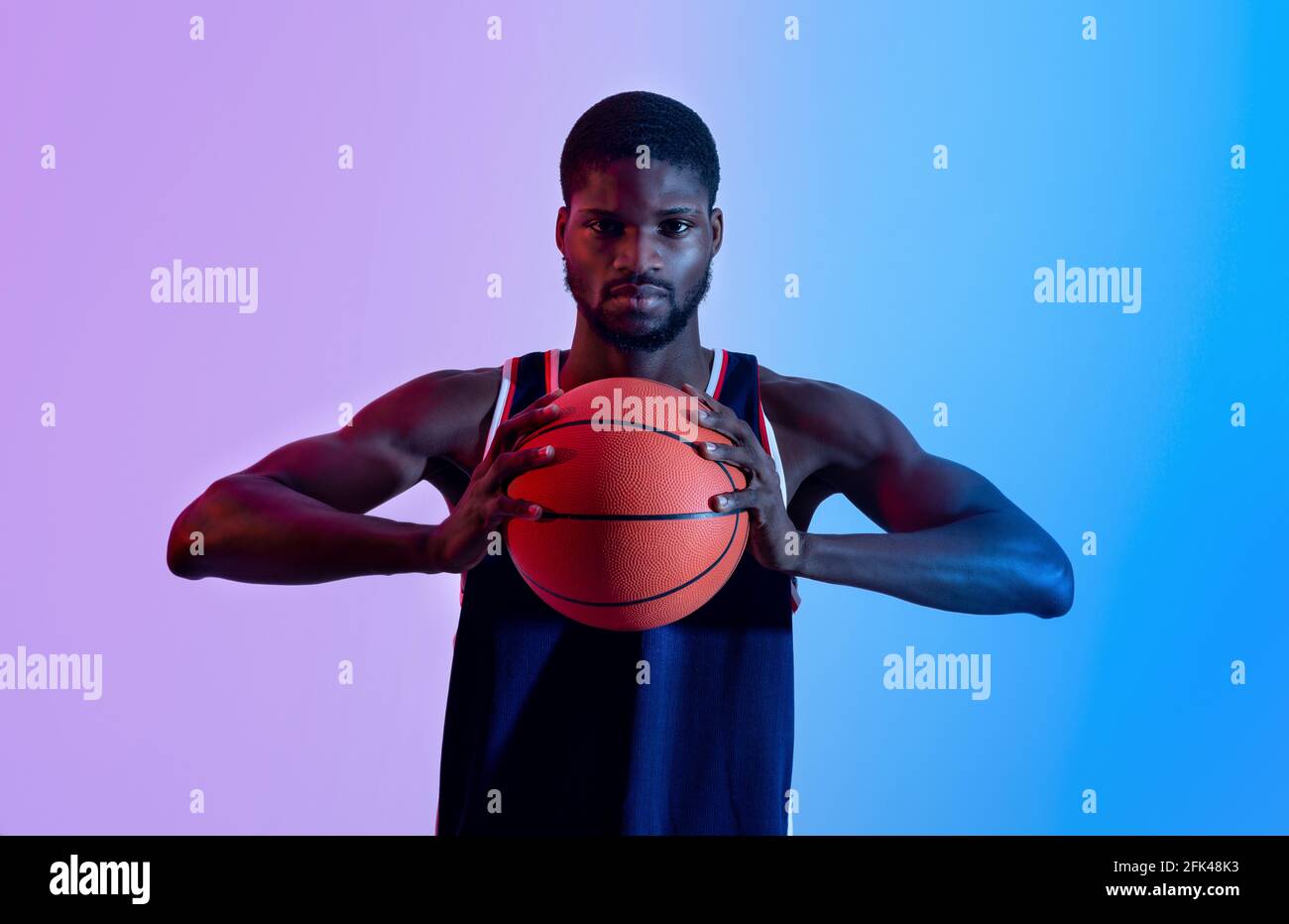 Ritratto di un giovane basketballer nero determinato che tiene la palla e guarda alla fotocamera in luce al neon Foto Stock