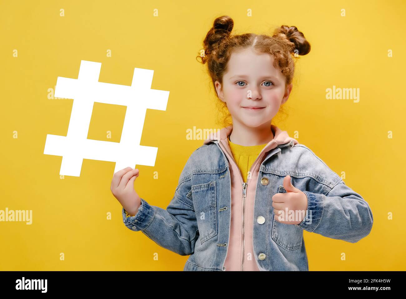 Carino caucasico ragazza bambino tenendo grande bianco hashtag simbolo che punta e mostra pollici verso l'alto gesto, indossa una giacca denim, isolato su parete gialla Foto Stock