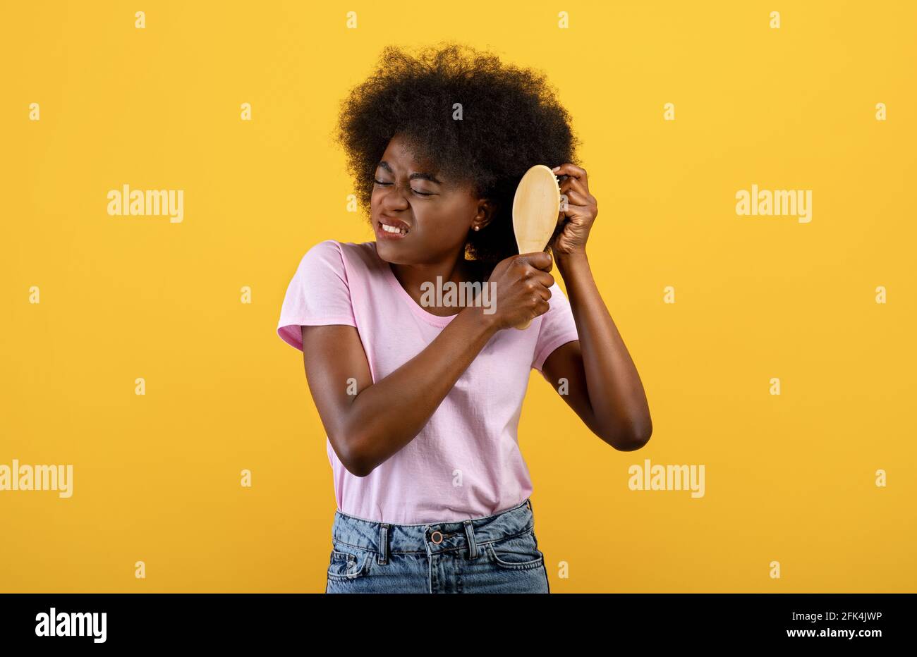 Infastidita donna afroamericana che ha problemi a spazzolare la sua riccia bush capelli su sfondo giallo studio Foto Stock